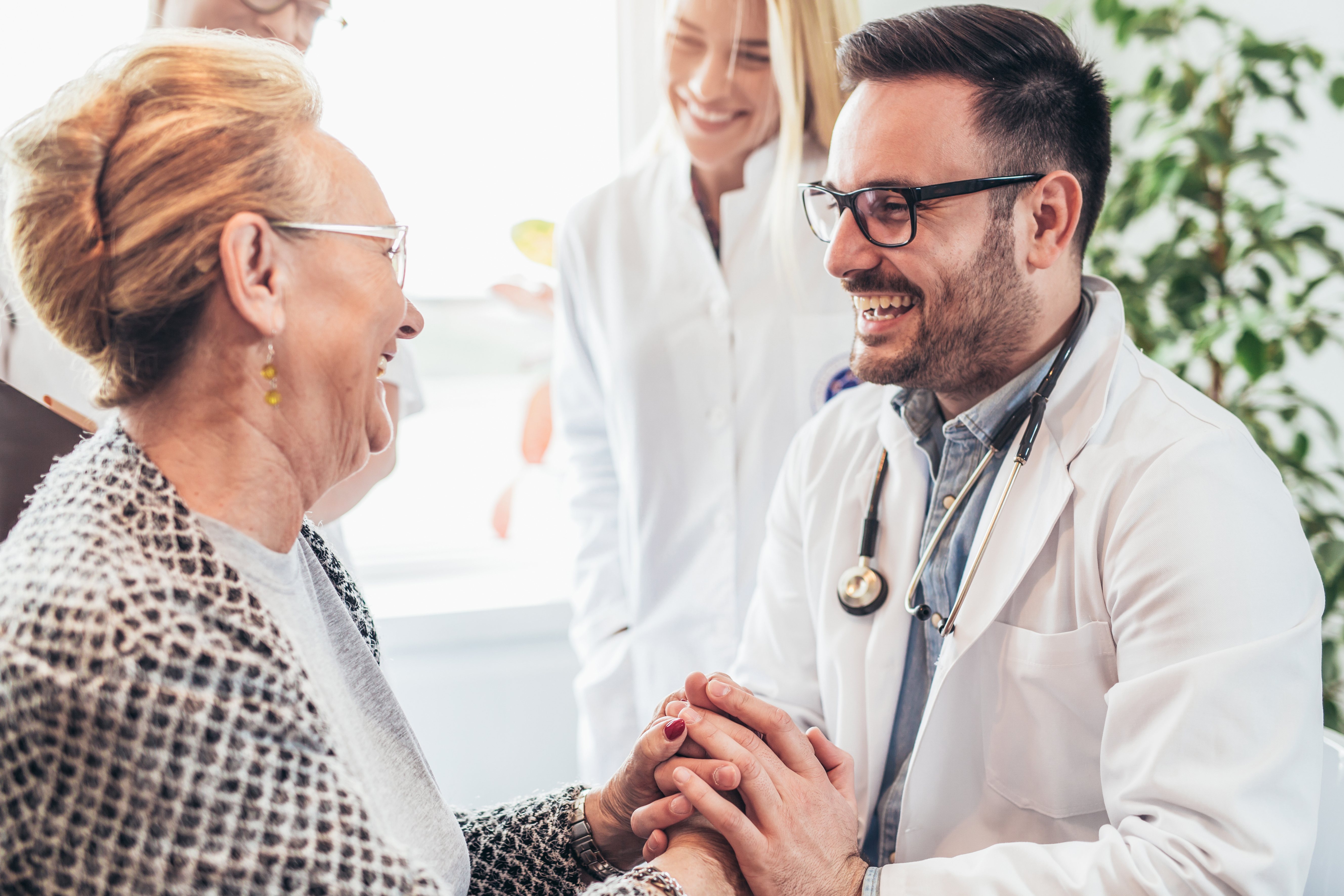 Ein Arzt hält die Hand einer Patientin. Beide lächeln sich an. Im Hintergrund stehen 2 weitere Personen.