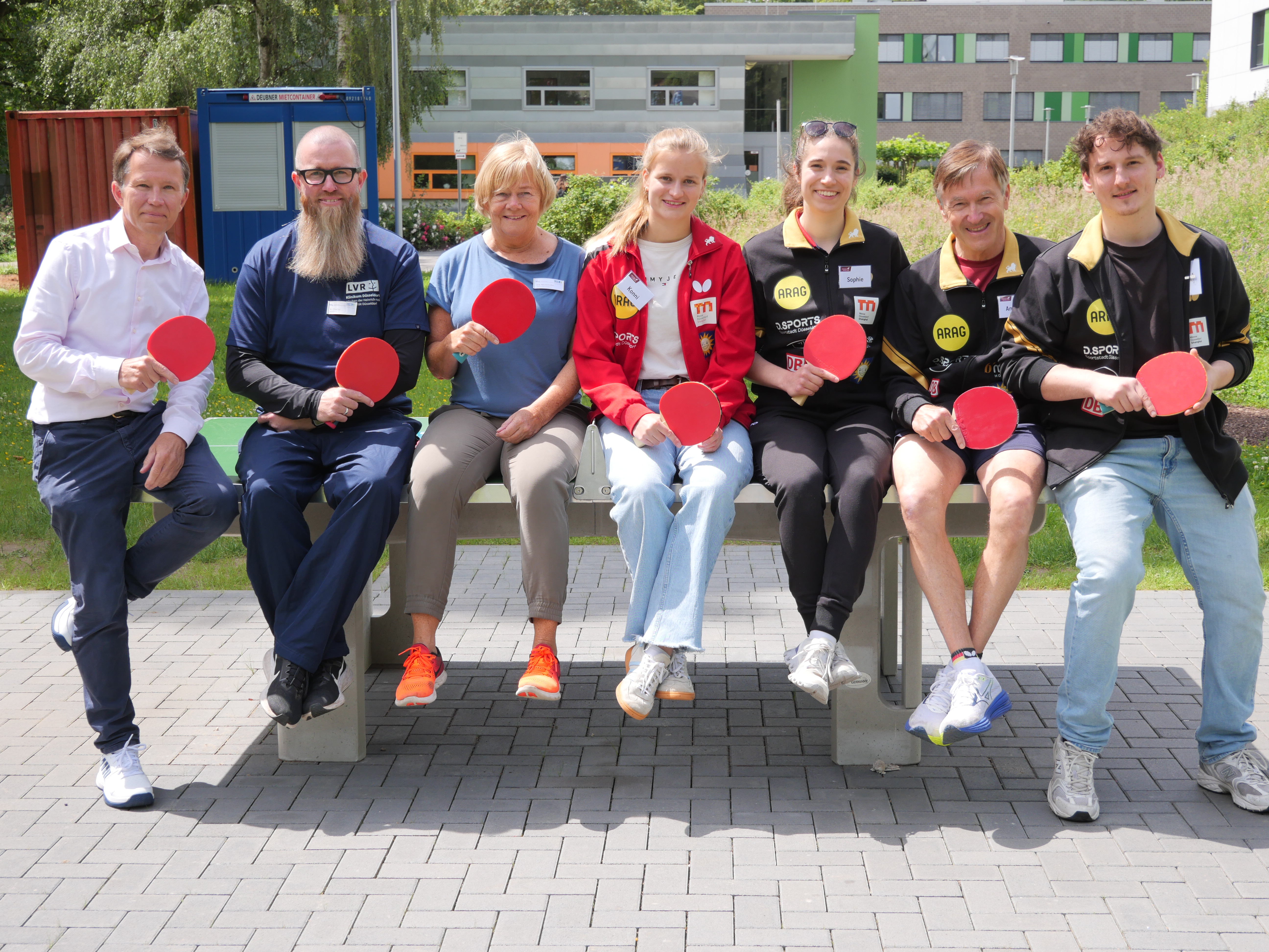 Auf dem Foto sind sieben Personen unterschiedlichen Alters und Geschlecht zu sehen. Sie sitzen auf einer Tischtennisplatte, halten Tischtennisschläger und schauen freundlich in die Kamera. 