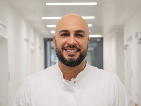 Portrait von Husni Khaled. Er trägt einen weißen Kittel und lächelt.