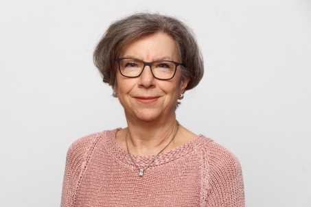 Eine Frau mit rosafarbenem Pullover und Brille lächelt vor einem weißen Hintergrund