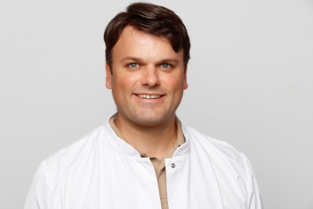 Ein Portrait von Dr. Faustmann. Ein Mann mit dunklen kurzen Haaren und einem weißen Kittel.