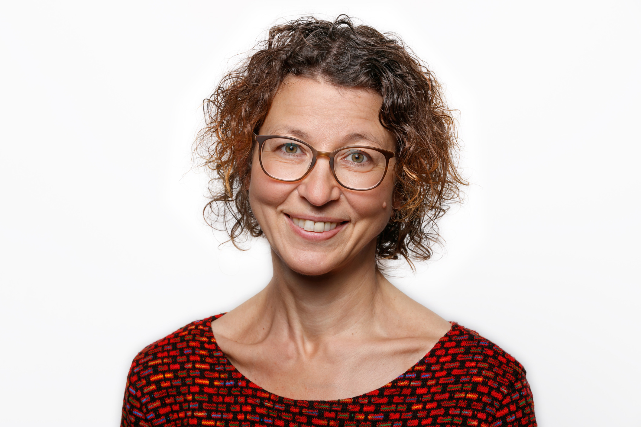 Ein Portrait einer Frau mit lockigen Haaren und einer Brille. Sie trägt ein rotes Oberteil.