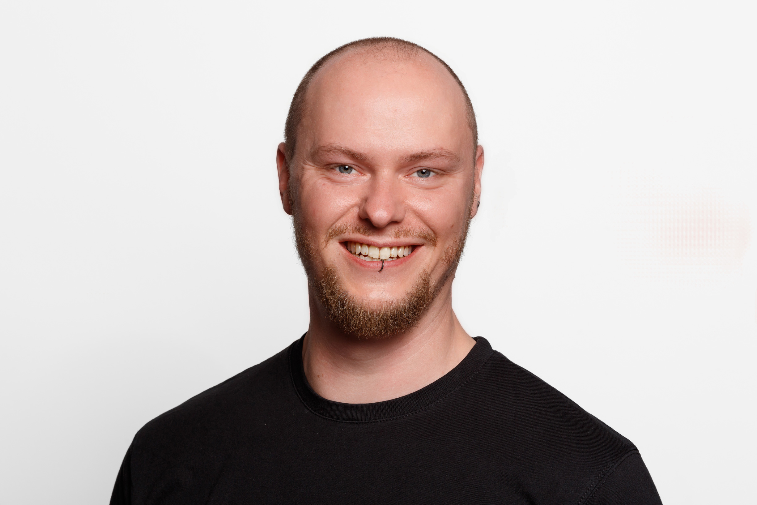 Portrait von einem Mann mit Piercing in der Unterlippe. Er trägt ein schwarzes T-Shirt und lächelt.