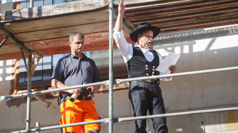 ein Zimmermann gibt den Richtspruch neben einem Mann mit orangener Arbeitshose 