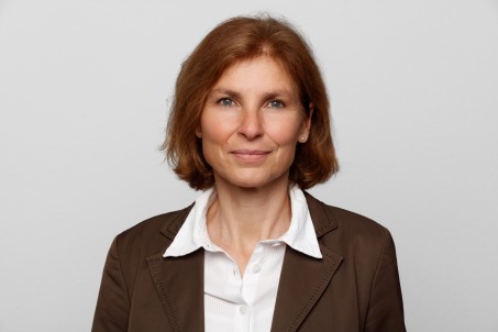 Zu sehen ist Frau Jörg. Eine Frau mit kinnlangen braunen Haaren und einer braunen Jacke. Sie lächelt und schaut nach vorne.