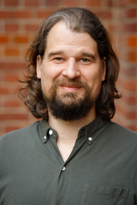 Dr. Fugmann mit Bart und schulterlangen Haaren vor einer Backsteinwand
