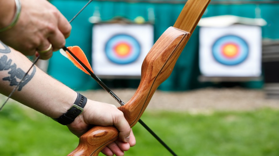 Arme beginnen einen Bogen zu spannen. Im Hintergrund stehen zwei Zielscheiben.