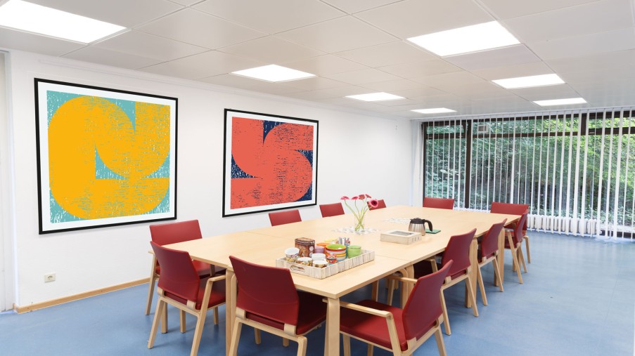 Ein großer Esstisch mit mehreren Stühlen steht in einem Raum mit einer Fensterfront, an der Wand hängen bunte Bilder