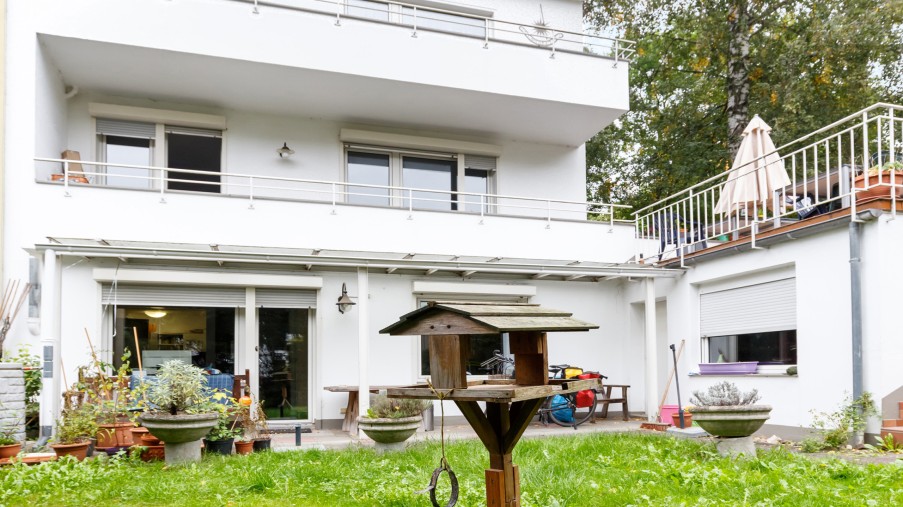 Ein Vogelhaus steht in einem Garten, dahinter sieht man die Rückseite des Hauses mit einer Terrasse und Balkonen