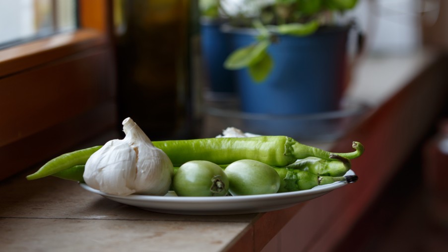 Eine Flasche Olivenöl, ein Topf mit Basilikum und Knoblauch, befinden sich auf einer Fensterbank