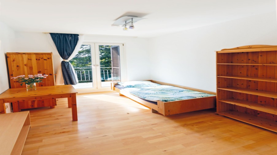 Raum mit einem Bett, Tisch, Regal und Kleiderschrank. Eine Glastür führt zum Balkon.