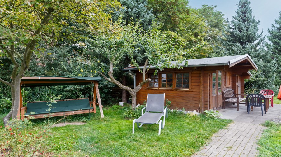 Eine Holzhütte steht in einem Garten, davor stehen Stühle und an der Seite steht eine Hollywoodschaukel