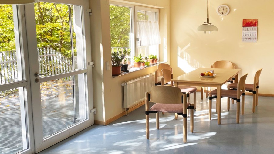 Ein Tisch mit Stühlen steht in einem Raum mit gelben Wänden, links daneben befindet sich eine Fensterfront